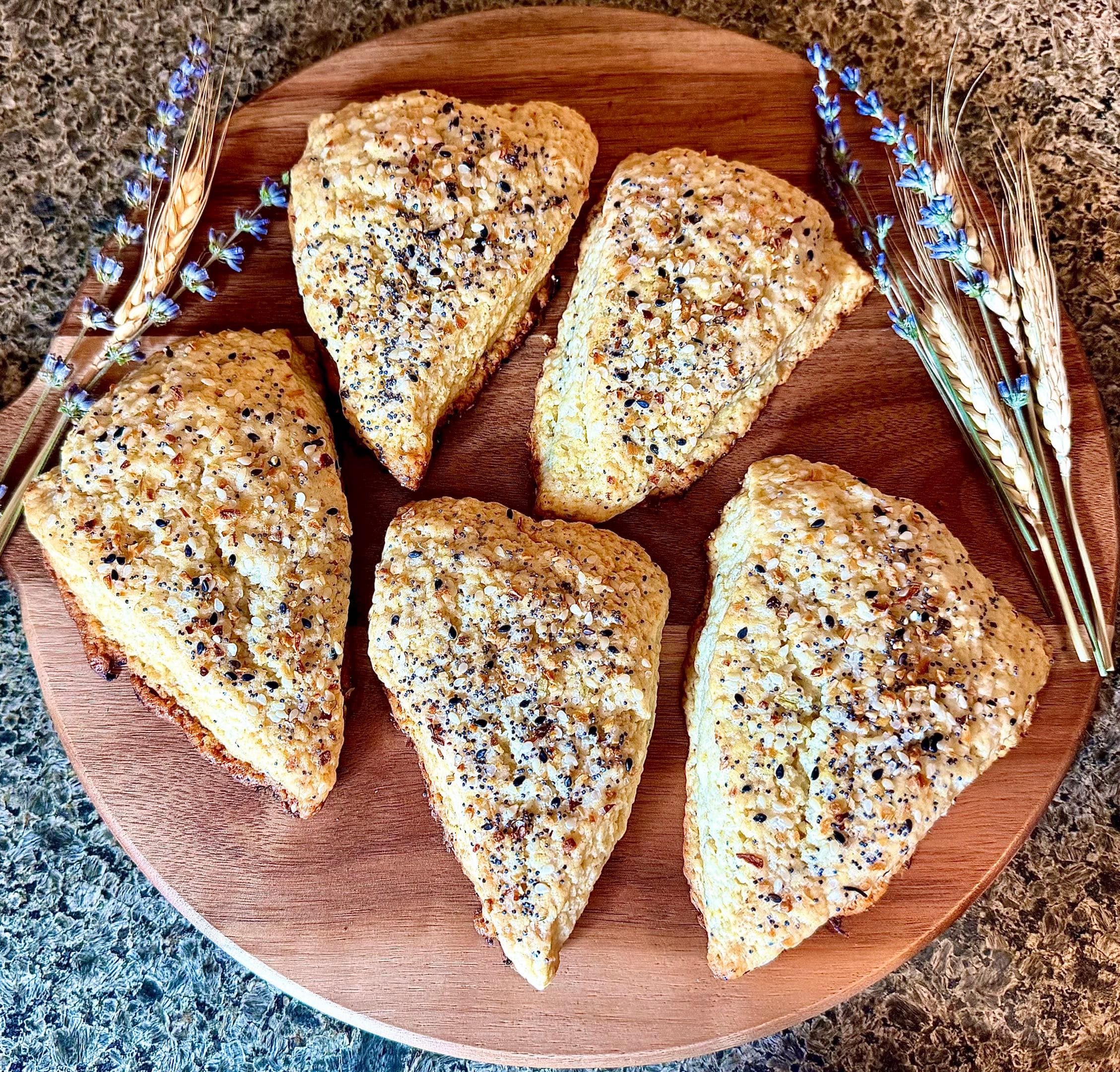 Six everything-seed scones on a round wooden board, framed by lavender sprigs and stalks of wheat